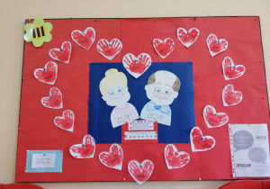 Na zdjęciu dekoracja na tablicy, na której widać wycięte z papieru kolorowe postacie babci i dziadka, otoczone w formie dużego serca małymi sercami wyciętymi z papieru i udekorowanymi odbitymi w farbie dłońmi dzieci ułożonych w kształcie serca.